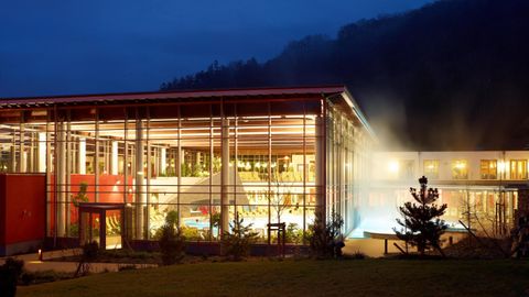 Een modern gebouw met grote ramen en een rood dak bij nacht. Op de achtergrond zijn bergen en dampend water te zien.