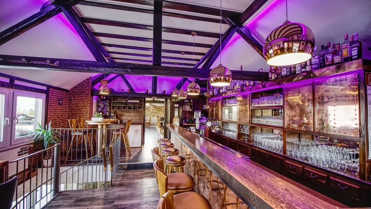 Eine moderne Bar mit einer langen Holztheke und stilvollen Beleuchtung. Die Wände sind in warmen Tönen gehalten, und der Raum hat eine einladende Atmosphäre.