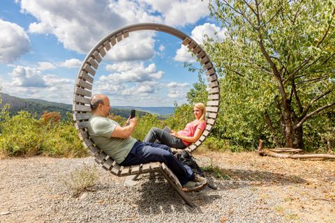 A couple is sitting relaxed on a round wooden bench outdoors. In the background, trees and a beautiful sky with clouds can be seen.