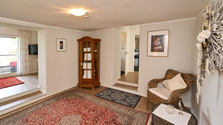 Een lichte woonkamer met een mooi tapijt en een houten kast. Er is een gezellige stoel en toegang tot andere delen van het huis.