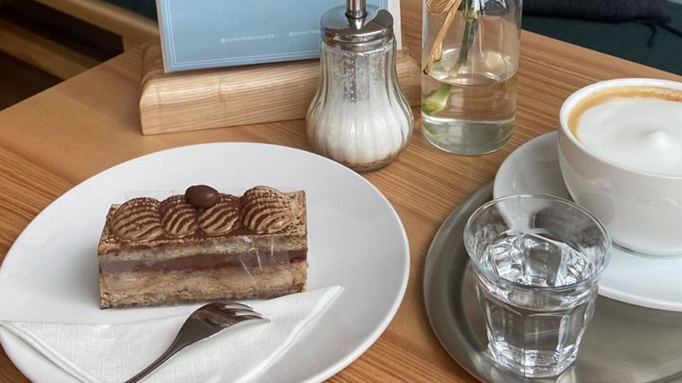 Een stuk taart op een bord, daarnaast een glas water en een kop koffie. Op de achtergrond zijn een suikerpot en een fles met water te zien.