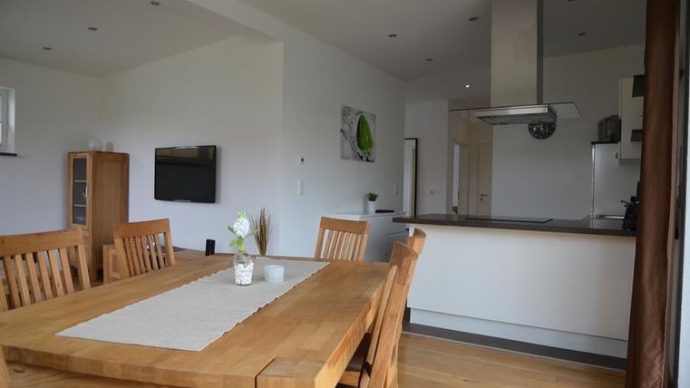 A bright and modern dining room with a large wooden table and chairs. In the background, an open kitchen can be seen.