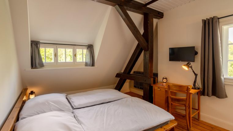 A bright room with a double bed and wooden flooring. There is a desk, a chair, and large windows that let in daylight.