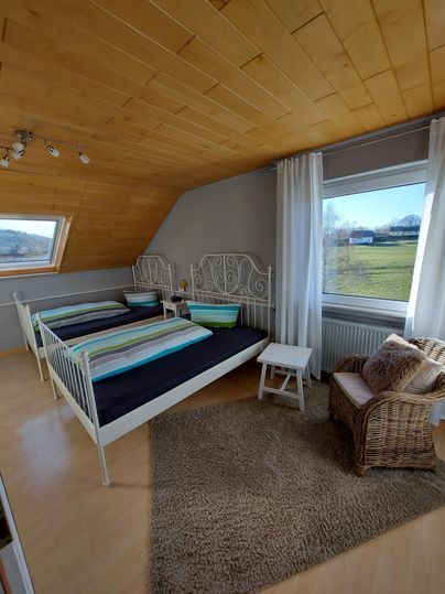 A cozy bedroom with two single beds and modern white furniture. Large windows provide a view of the green landscape.
