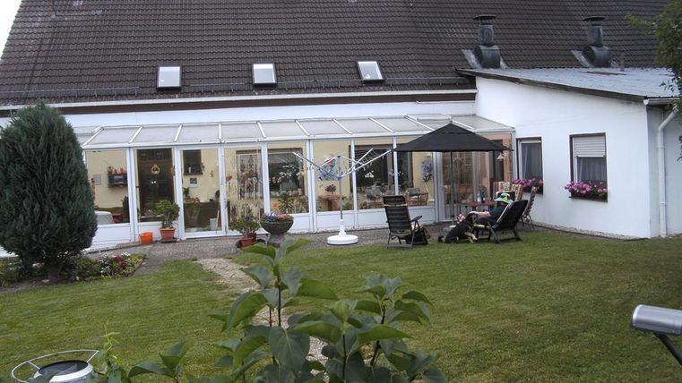 Ein kleiner Garten mit einer Terrasse und einem Gewächshaus. Im Hintergrund sieht man ein zweigeschossiges Haus und einige Pflanzen.