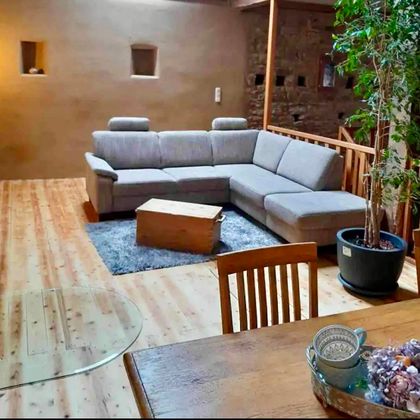 Ein gemütliches Wohnzimmer mit einer grauen Couch und einem Holztisch. Pflanzen und ein Holzfußboden sorgen für eine einladende Atmosphäre.