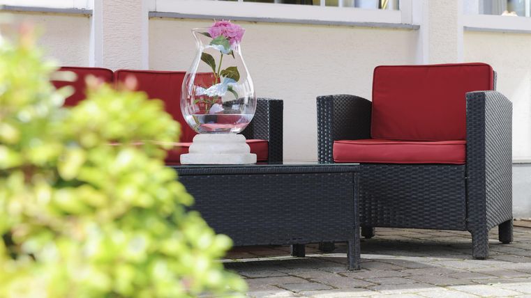 Ein gemütlicher Sitzbereich mit roten Stühlen und einem Tisch in einem hellen Außenbereich. Eine Vase mit einer Blume dekoriert den Tisch.