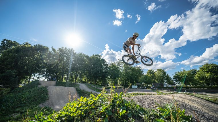 Ein BMX-Fahrer springt über eine Rampe in einem Park. Die Sonne scheint hell am Himmel, umgeben von Bäumen und Wolken.