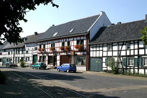 Eine malerische Straße mit traditionellen Fachwerkhäusern. Auf beiden Seiten stehen einige Autos und es ist schönes Wetter.
