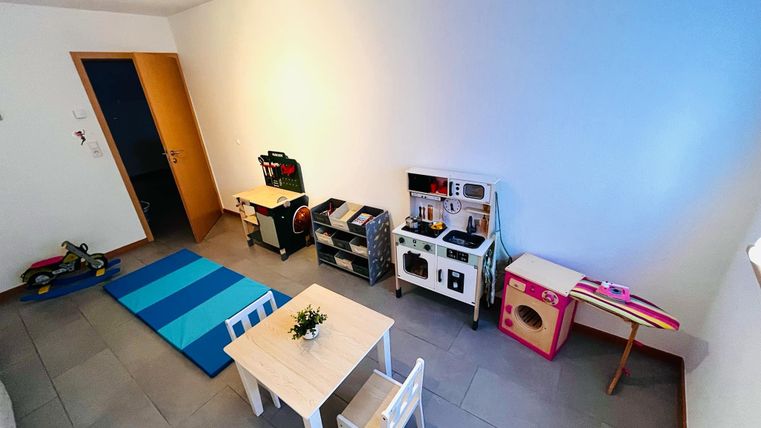 A playroom with a small table and chairs. Against the wall are kitchen and play equipment, and a blue rug is on the floor.