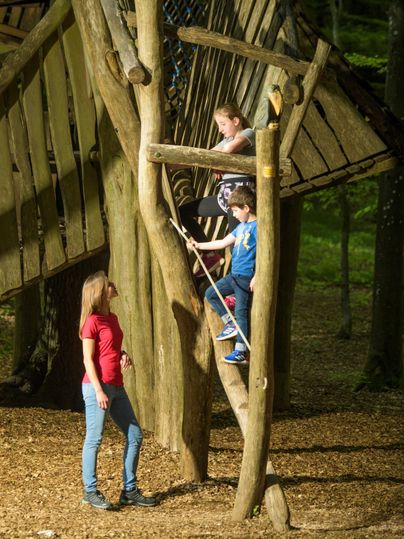 Kinder spielen auf einem Klettergerüst aus Holz im Wald. Eine Erwachsene beobachtet sie lächelnd.