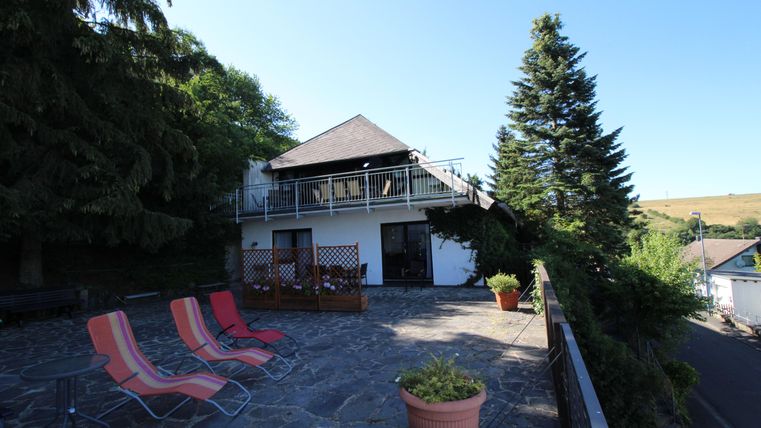 Ein modernes Haus mit einem großzügigen Außenbereich. Rote Liegestühle stehen auf der Steinoberfläche, umgeben von Bäumen und Pflanzen.