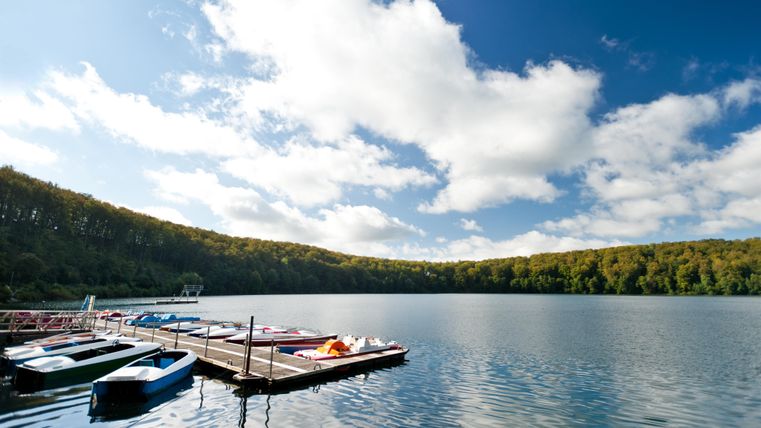 Ein ruhiger See mit Booten an einem Steg. Der Himmel ist blau mit einigen Wolken und die Ufer sind mit Bäumen gesäumt.
