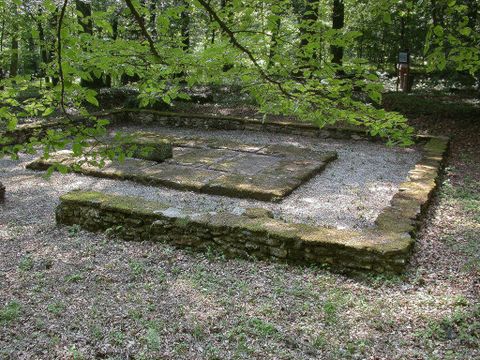 A historical ruin in a wooded area, surrounded by green landscapes. The stones are partially covered with moss and the ground is covered with gravel.