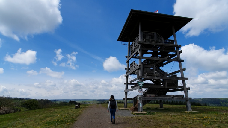 Eine Person geht auf einem Weg zu einem Aussichtsturm. Der Himmel ist blau mit vielen Wolken und die Landschaft ist grün.