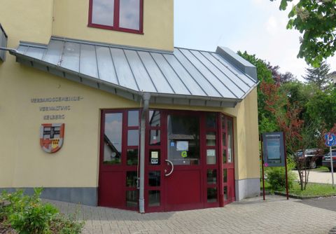 Ein modernes Gebäude mit einem Eingangsbereich aus Glas und roten Türen. Im Hintergrund sind einige Bäume und eine beschilderte Umgebung sichtbar.