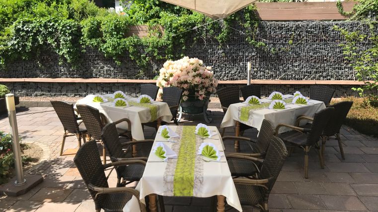 Ein gemütlicher Außenbereich mit mehreren Tischen und Stühlen. Auf den Tischen liegen helle Tischdecken mit grünen Mustern und eine große Blumenvase ist im Hintergrund zu sehen.
