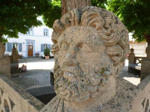 Eine antike Steinbüste mit einer detaillierten Darstellung eines bärtigen Mannes. Im Hintergrund sind grüne Bäume und ein historisches Gebäude sichtbar.