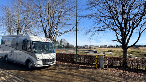 Ein Wohnmobil steht auf einem Parkplatz zwischen Bäumen. Im Hintergrund ist eine weitläufige Wiese zu sehen.