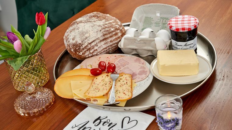 A breakfast dish on a tray with bread, cheese, ham, tomatoes, and eggs. A flower and a candle are placed on the table as well.