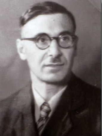 A black-and-white portrait of a man wearing glasses and a suit. He is looking at the camera and has a neutral expression.