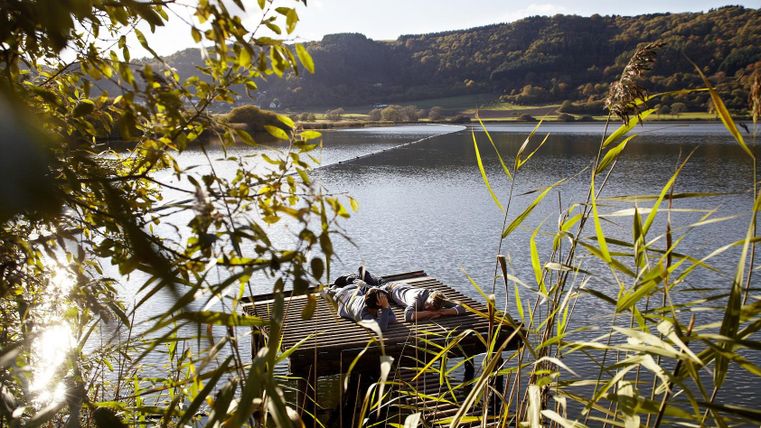 Ein kleiner Steg am Wasser, umgeben von grünen Pflanzen. Im Hintergrund sind sanfte Hügel und ein klarer Himmel zu sehen.