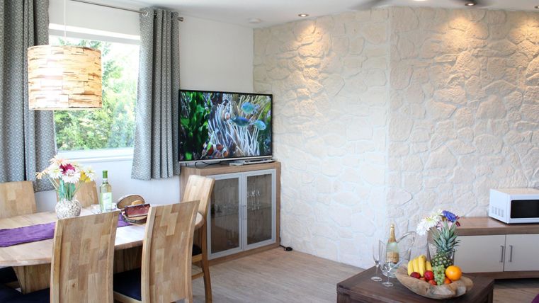 Ein modernes Wohnzimmer mit einem Essbereich aus Holz. Ein Fernseher und eine dekorative Obstschale sorgen für eine gemütliche Atmosphäre.