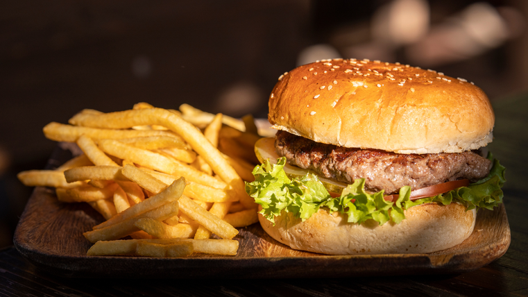Een sappige burger met sesambroodje, sla en tomaat ligt op een houten plank. Naast hem liggen knapperige frietjes.
