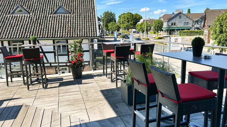 Een modern terras met tafels en stoelen in een restaurant. Op de achtergrond zijn huizen en bomen te zien, onder een heldere blauwe lucht.