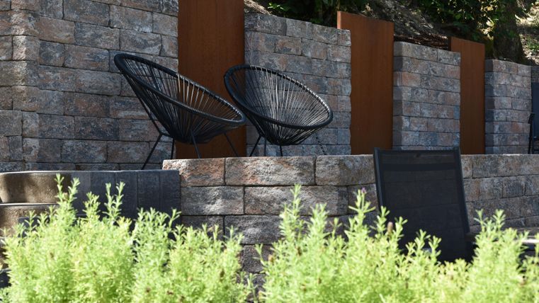 Zwei schwarze Korbstühle stehen vor einer Steinmauer. Grüne Pflanzen im Vordergrund schaffen eine einladende Atmosphäre.