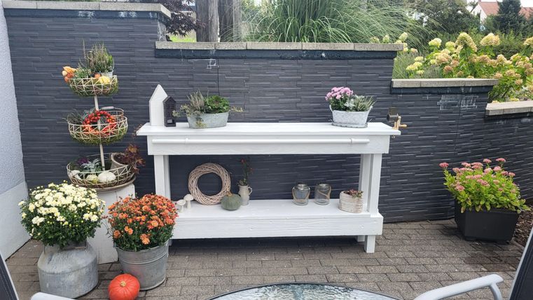 A beautiful garden table with several plant pots and containers. Surrounded by colorful flowers and a modern wall.