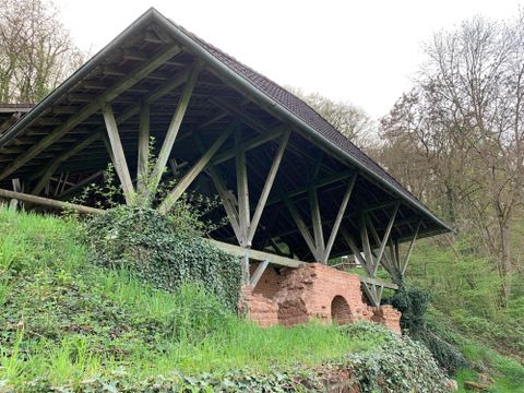 Eine historische Struktur mit einem Holzgestell, die an einem Hang steht. Die Umgebung ist von grünem Gras und Bäumen umgeben.