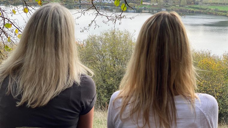 Two women are sitting on a bench and gazing at a calm lake. In the background, trees and hills can be seen.
