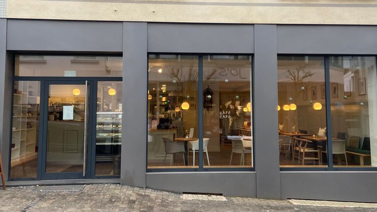 Een modern café met grote ramen en een uitnodigende interieur. Buiten staat een bord dat gasten aantrekt.