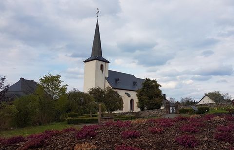 Eine kleine Kirche mit einem hohen Turm steht neben einem Blumenbeet. Der Himmel ist bewölkt und die Umgebung ist grün.