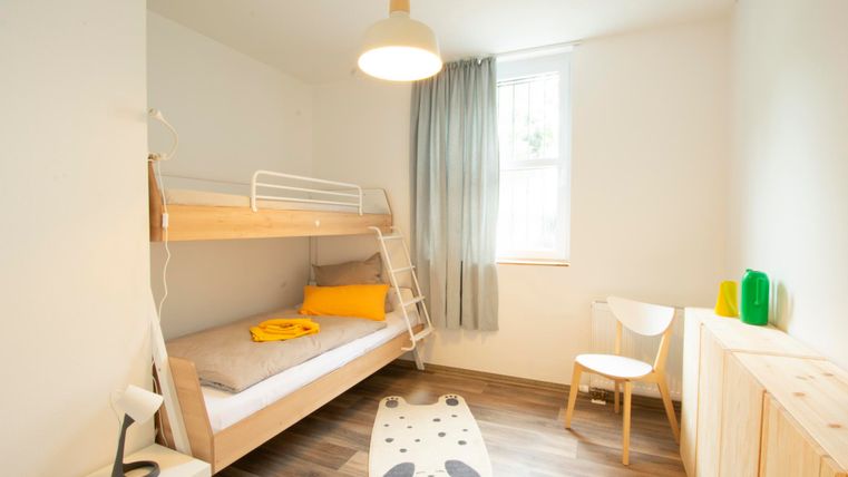 Ein gemütliches Schlafzimmer mit Hochbett, einem Stuhl und einer hellen Fensterfront. Die Farben sind freundlich und der Raum wirkt einladend.