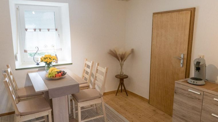 Ein helles Zimmer mit einem Holztisch und vier Stühlen. Auf dem Tisch steht eine Obstschale und eine Vase mit Sonnenblumen.