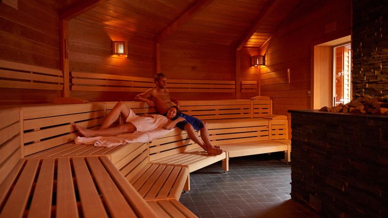Een gezellige sauna-ruimte met houten planken en zachte verlichting. Twee mensen ontspannen zich op de banken.