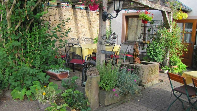 Ein gemütlicher Außenbereich mit Tischen und Stühlen, umgeben von Pflanzen und Blumen. Die Atmosphäre ist einladend und freundlich.