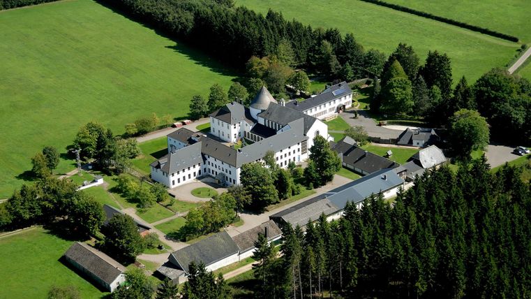 Ein großes Gebäude umgeben von grünen Feldern und Wäldern. Die Ansicht ist aus der Vogelperspektive und zeigt eine ruhige ländliche Umgebung.