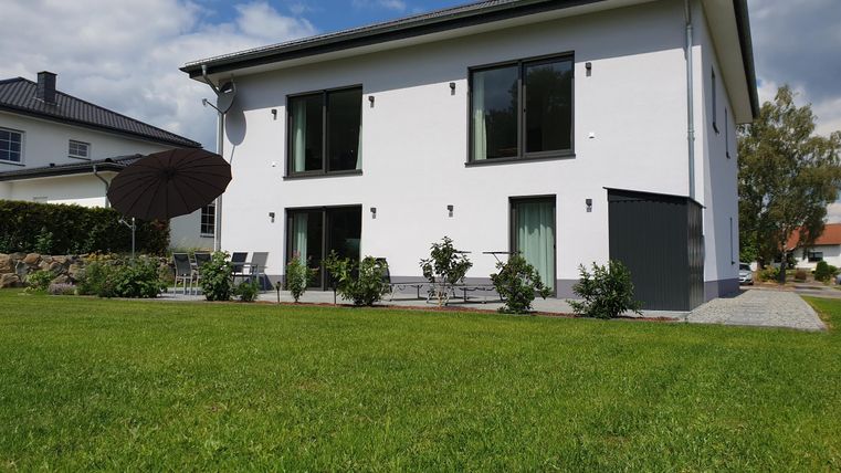 Een modern, wit huis met grote ramen staat op een groene weide. Op de achtergrond zijn enkele bomen en een blauwe lucht te zien.