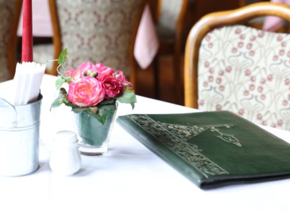 An elegant table in a restaurant with a green menu and a vase with pink flowers. In the background, decorated chairs can be seen.
