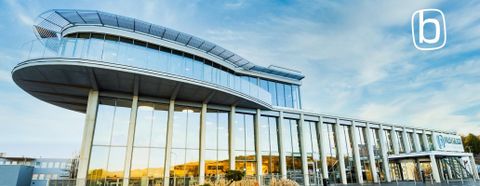 Ein modernes Gebäude mit einer großflächigen Glasfassade und einem auffälligen Dach. Der Himmel ist blau und leicht bewölkt.