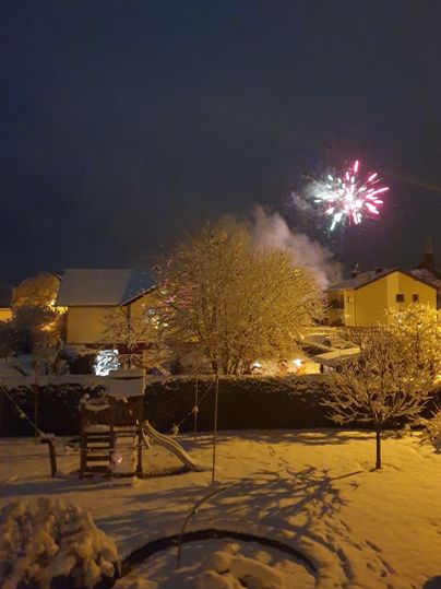 Eine verschneite Winterlandschaft mit bunten Feuerwerken am Nachthimmel. Die Häuser sind festlich beleuchtet und die Bäume sind mit Schnee bedeckt.