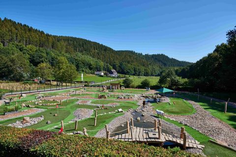 A spacious nature playground in a green environment. The sky is clear and blue, surrounded by gentle hills and trees.