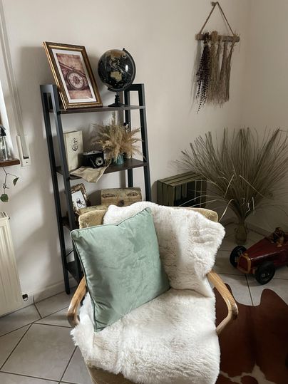 Een gezellige ruimte met een stoel, die is uitgerust met een warme deken en een kussen. Op de achtergrond staat een boekenplank met decoratie-elementen en een plant.