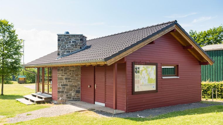 Ein rotes Haus mit einem Steinsockel und einem schrägen Dach. Es liegt in einer grünen Umgebung mit klarem Himmel.