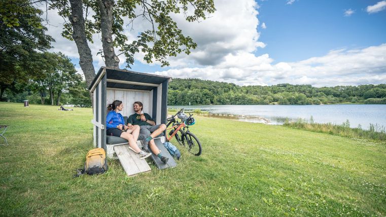 Zwei Personen sitzen in einem kleinen Unterstand am See und genießen die Aussicht. Neben ihnen steht ein Fahrrad auf dem grünen Rasen.