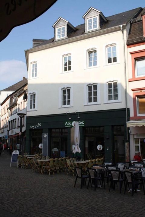 Een licht gebouw met verschillende ramen en een restaurant op de begane grond. Voor het restaurant staan tafels en stoelen op een geplaveide straat.
