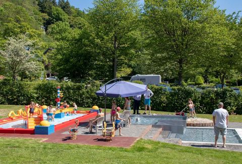 Ein schöner Garten mit einem Planschbecken für Kinder und bunten Spielzeugen. Im Hintergrund sind grüne Bäume und einige Erwachsene zu sehen, die entspannen.